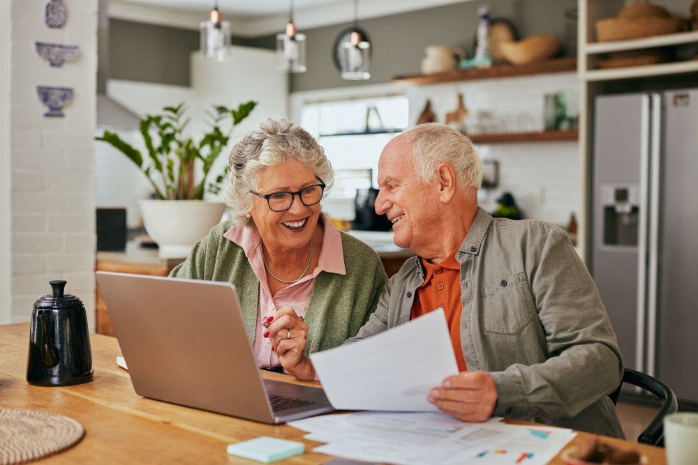 Elderly couple look happy, both are looking at a laptop and paperwork in their kitchen